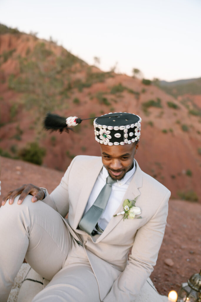 A man in a light beige suit and gray tie sits outdoors on a rocky hillside, wearing a traditional decorated hat with beads and a feather. He is smiling and has a white boutonniere pinned to his jacket.