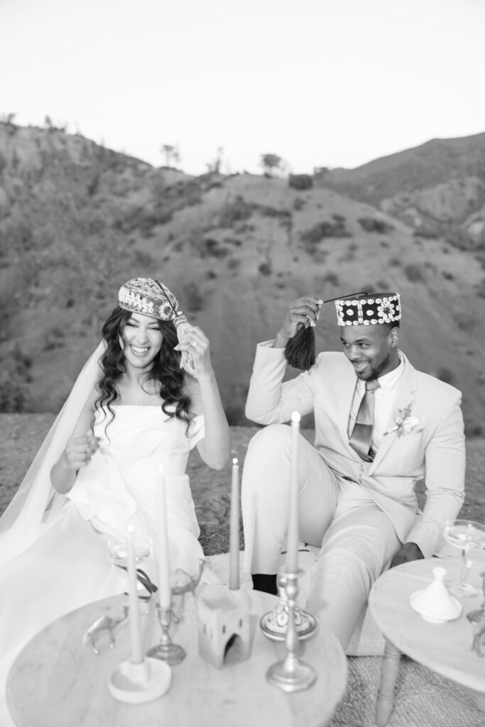A bride and groom sit outdoors, smiling and placing ornate hats on their heads. They are dressed in wedding attire, with mountains in the background and decorative candles and objects on the tables in front of them.