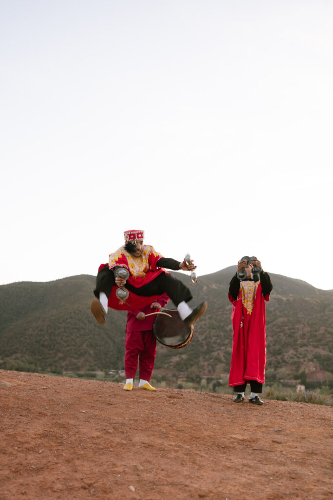Two people in traditional red clothing and masks stand on a dirt hill; one is mid-jump with legs spread, holding silver objects, while the other watches. Green hills and a pale sky are in the background.