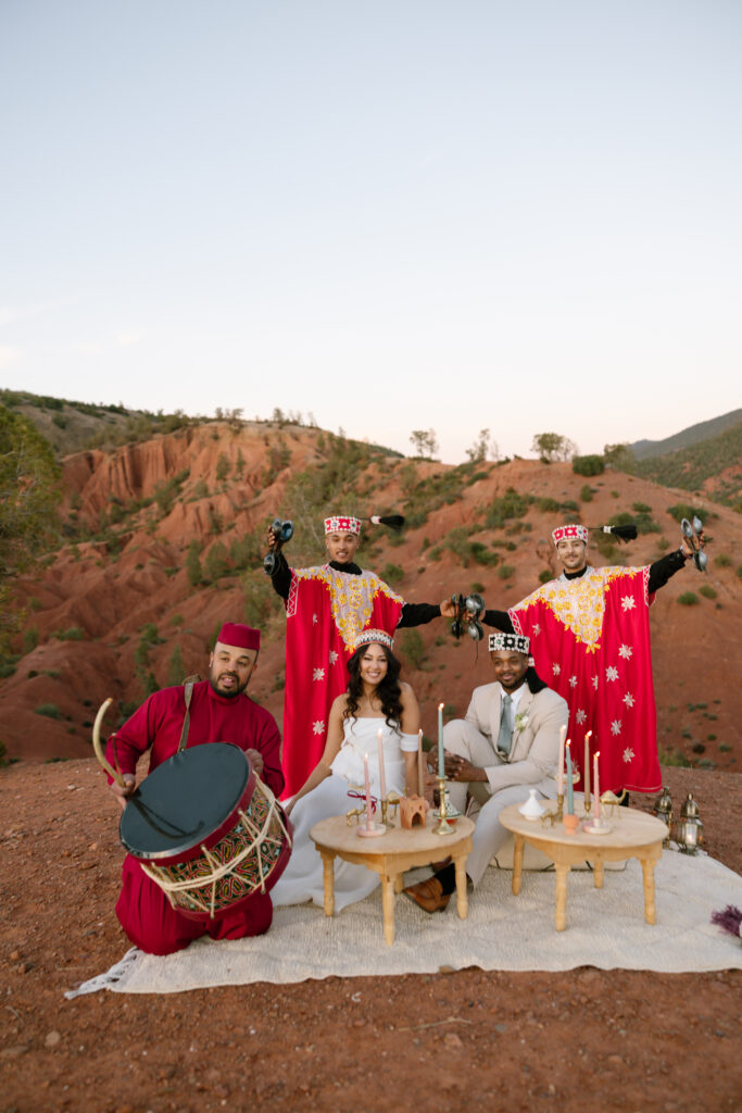 A couple sits at a picnic setup with low tables, food, and candles on a rug outdoors, while a drummer in red and three performers in elaborate red and gold costumes and masks stand behind them with arms raised against a scenic rocky landscape.