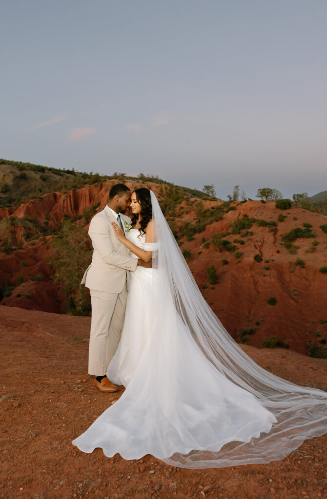 A bride and groom embrace on a red rocky landscape at sunset. The bride wears a long white gown with a flowing veil, and the groom is dressed in a light beige suit. They gaze lovingly at each other.
