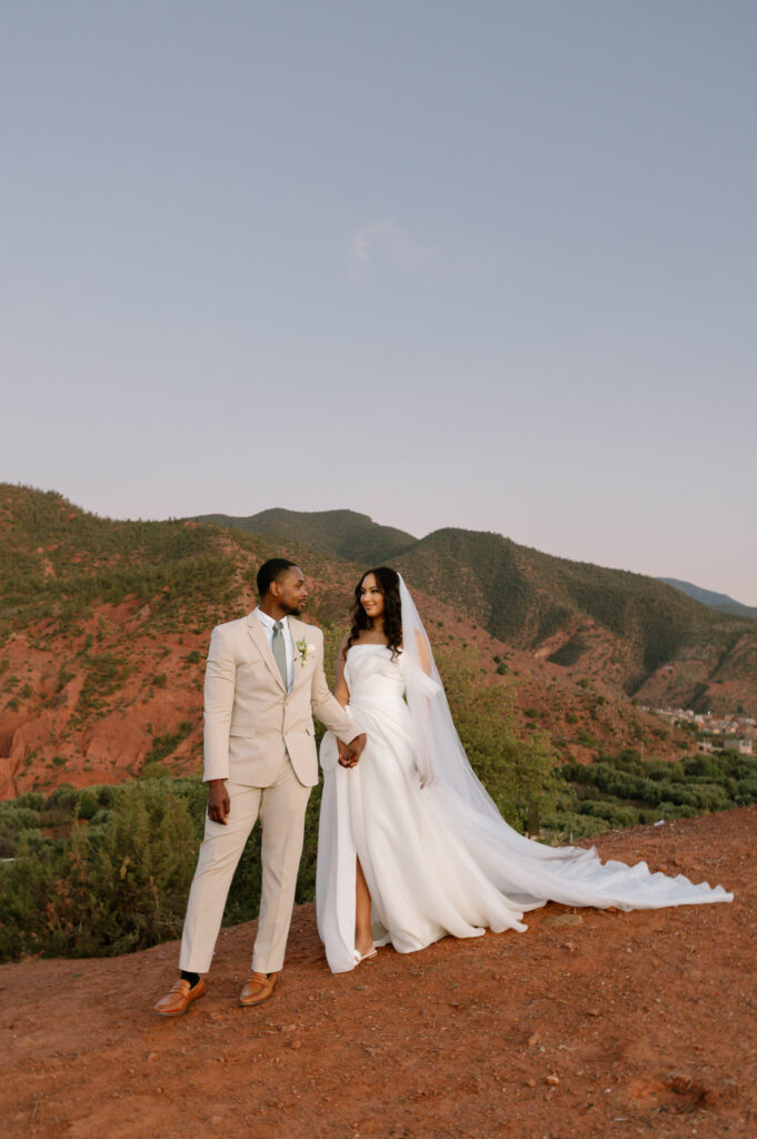 A bride in a white gown and long veil stands holding hands with a groom in a beige suit and blue tie. They are outdoors on reddish terrain with green hills and a clear sky in the background.