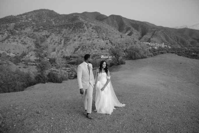 A bride and groom walk hand in hand on a gravel path with mountains in the background. The scene is peaceful and outdoors, captured in black and white.
