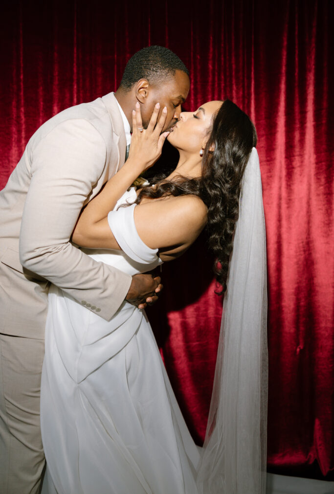 A bride and groom share a romantic kiss in front of a red velvet curtain. The groom wears a beige suit, and the bride wears an off-shoulder white dress with a long veil. They are embraced in a joyful, intimate moment.