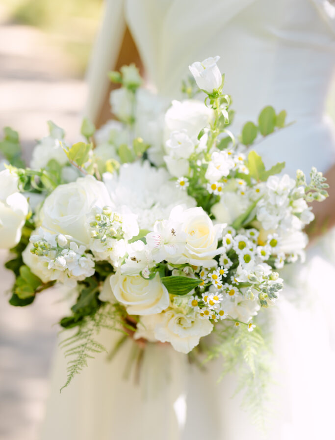 A bride in a white dress holds a bouquet of white roses, daisies, and greenery, with sunlight softly illuminating the flowers.