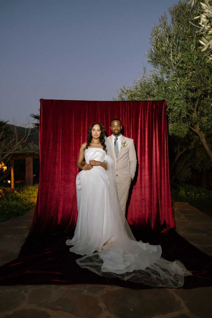 A bride in a white gown and veil stands beside a groom in a light suit and tie. They pose together in front of a deep red velvet backdrop outdoors at dusk, surrounded by trees and greenery.