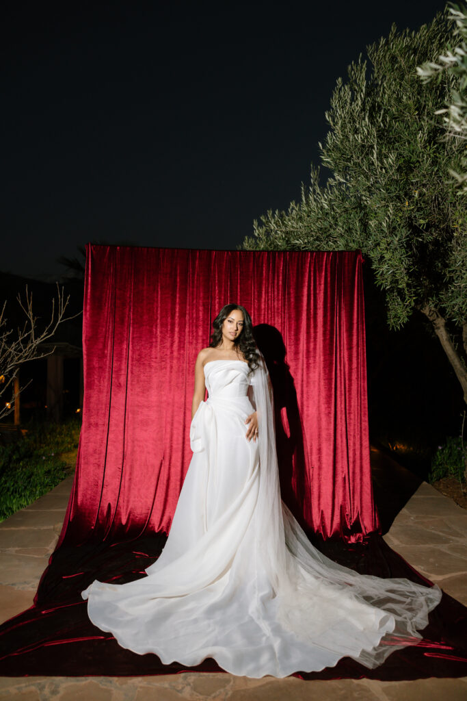 A woman in a strapless white wedding gown stands in front of a rich red velvet backdrop outdoors at night, with greenery and trees visible at the edges of the scene.