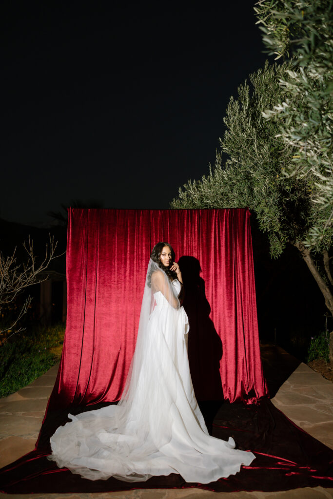A bride in a flowing white gown and veil stands on a stone path in front of a red velvet backdrop at night, surrounded by greenery and trees.