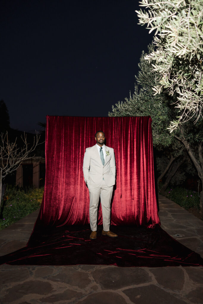 A man in a light gray suit and green tie stands confidently on a stone path in front of a red velvet curtain backdrop, with trees and plants surrounding him in an outdoor nighttime setting.