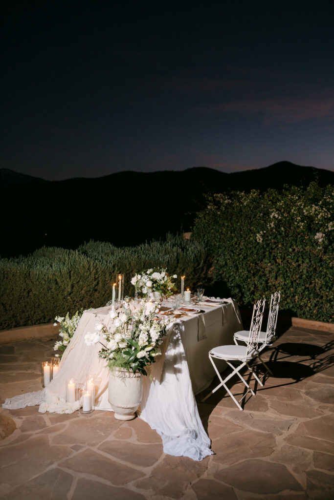 A romantic outdoor table for two is elegantly set with white flowers, candles, and flowing white fabric, surrounded by greenery and mountains at dusk.