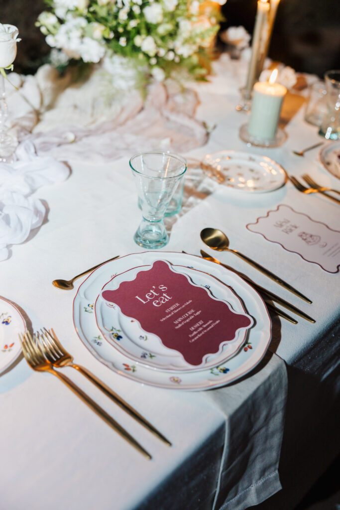 A formal table setting with gold utensils, glassware, floral china plates, and a maroon menu card reading Lets Eat placed on a white tablecloth, with candles and flowers in the background.
