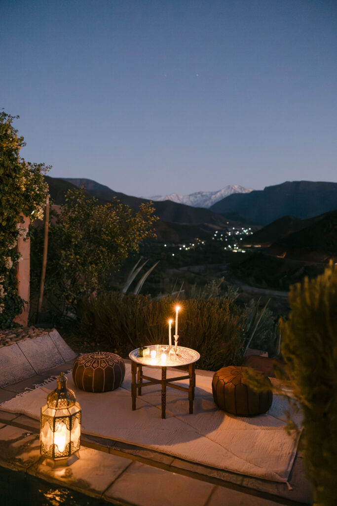 A cozy outdoor setting at dusk features a low table with lit candles, two poufs, a white rug, and a decorative lantern, overlooking mountains and distant village lights under a twilight sky.