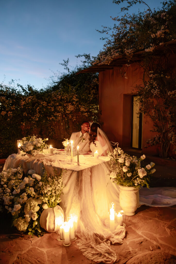 A bride and groom sit at a candlelit table decorated with white flowers and draped fabric during an outdoor evening wedding, sharing an intimate moment under soft, warm lighting.