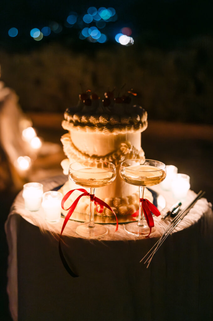 A white cake decorated with cherries and cream sits on a table surrounded by lit candles. In front of the cake are two champagne glasses tied with red ribbons, set against a dark, softly blurred background.