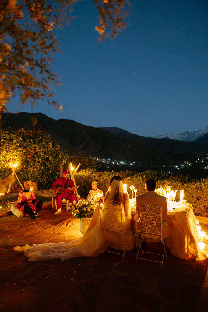 A bride and groom sit at a candlelit outdoor table at night, facing a mountain landscape. Musicians in vibrant attire play traditional instruments nearby under a dark, starry sky.