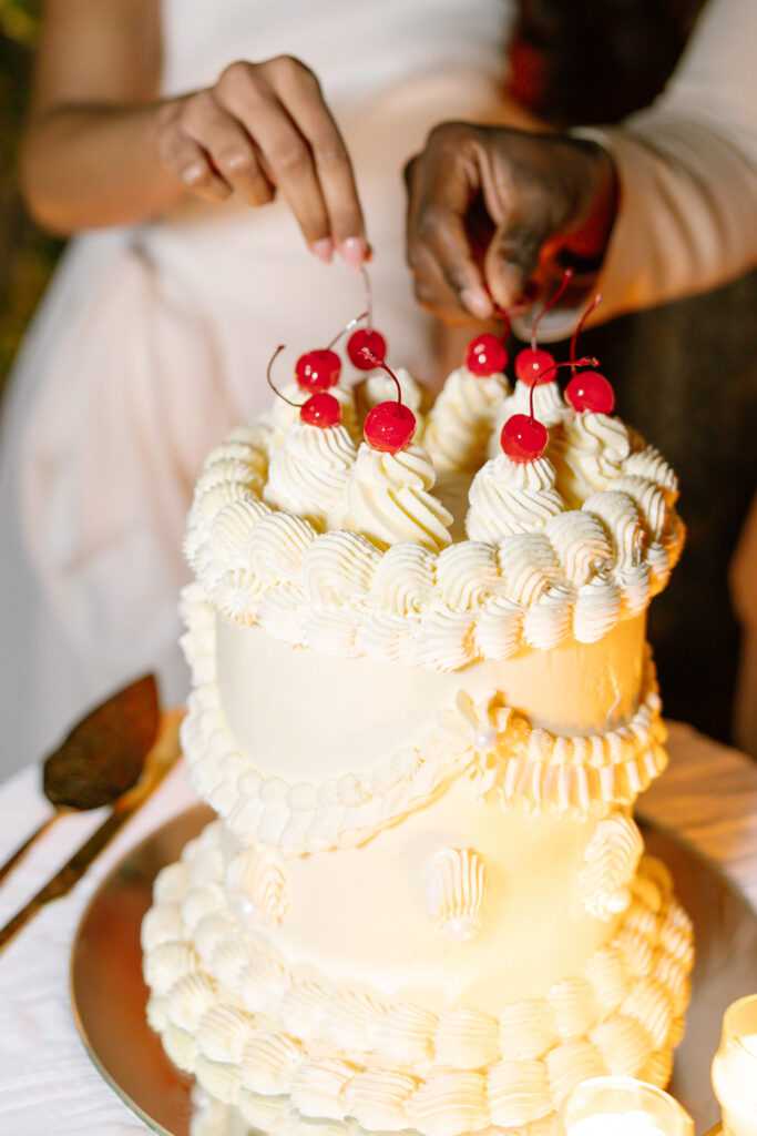 Two people decorate a white, double-tiered cake with swirls of frosting and red cherries. The cake is on a silver tray, and one person is wearing white while the other has a darker skin tone.
