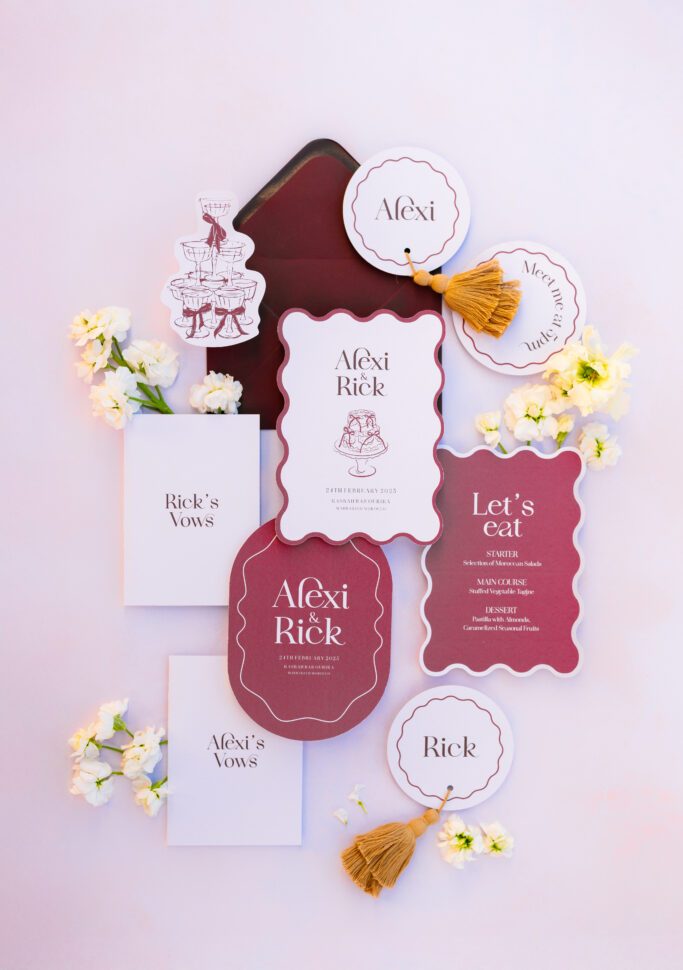 A flat lay of elegant wedding stationery in burgundy, white, and gold tones, including invitations, vow booklets, menus, place cards, and paper tassels, arranged with small white flowers on a pale pink background.