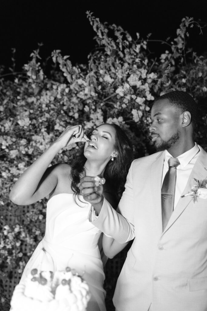 A bride in a strapless dress smiles as she feeds herself cake, while the groom in a light suit holds her hand and looks at her. They stand outdoors in front of a backdrop of flowers at night.