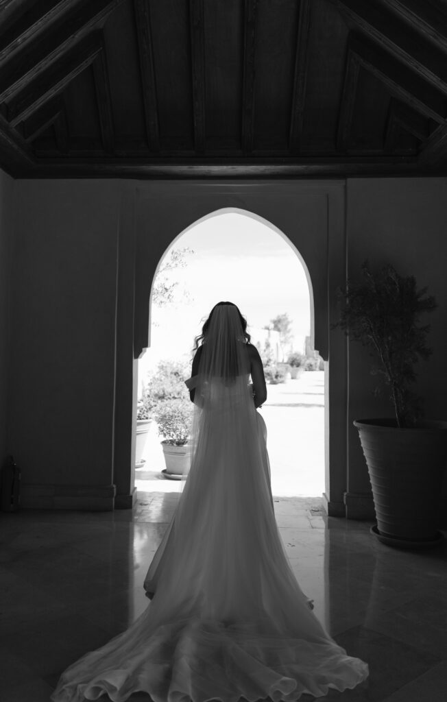 A bride in a flowing wedding dress and veil stands inside under a wooden ceiling, facing an arched doorway that opens to a sunlit outdoor scene. The image is in black and white.