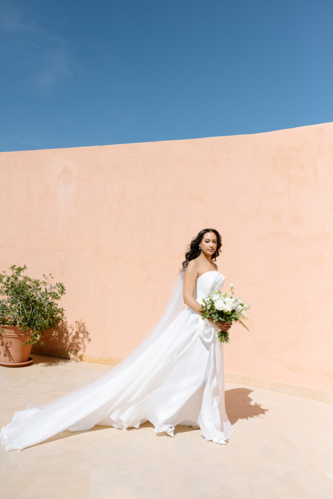 A bride in a flowing white gown holds a bouquet of white and green flowers, standing outdoors against a peach-colored wall and clear blue sky, with a potted plant in the background.