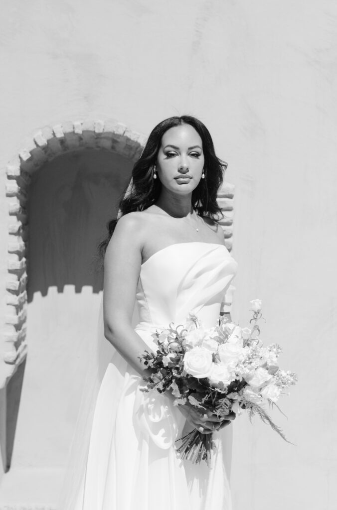 A bride in a strapless white gown holds a bouquet of white flowers, standing in front of a textured wall with an arched alcove, looking calmly at the camera. The image is in black and white.