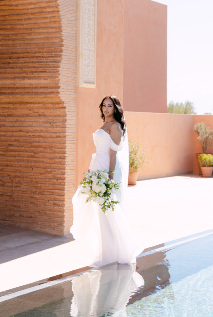 A bride in a flowing white dress holding a bouquet of white and green flowers stands beside a pool, with a peach-colored building and potted plants in the background.