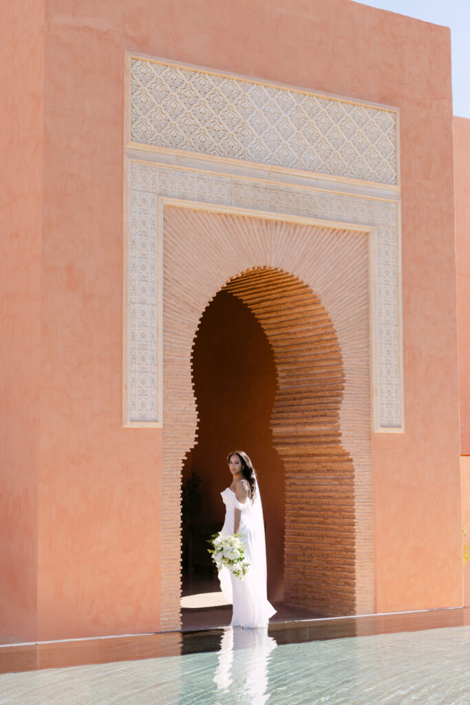 A bride in a white dress holding a bouquet stands in front of a large, ornate arched doorway with intricate patterns, reflected in a pool of water at the base of a terracotta-colored building.