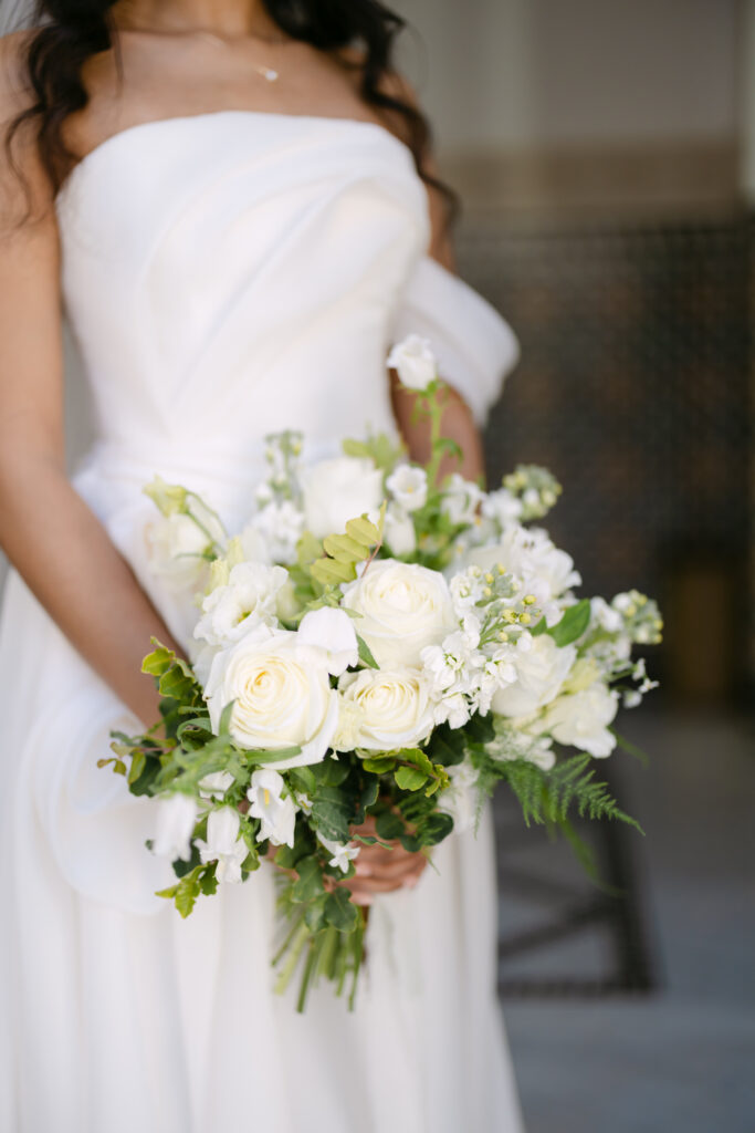 A bride in a strapless white dress holds a bouquet of white roses and greenery. Her long dark hair falls over her shoulders, and the background is softly blurred.