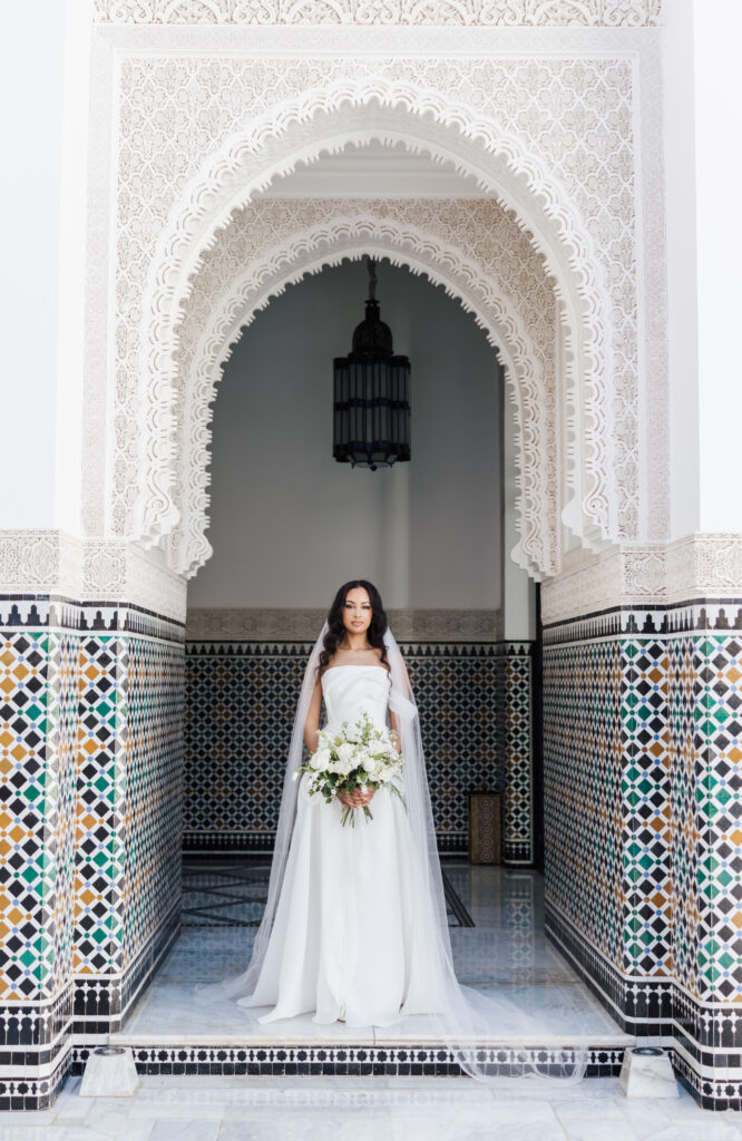 A bride in a white dress and veil stands under an ornate, arched doorway with intricate carvings and colorful geometric tilework, holding a bouquet of white flowers.