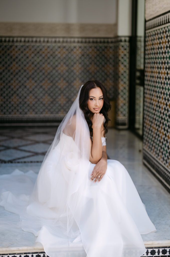 A bride in a white gown and veil sits on marble steps, resting her chin on her hand, with patterned tile walls in the background. She gazes thoughtfully at the camera.
