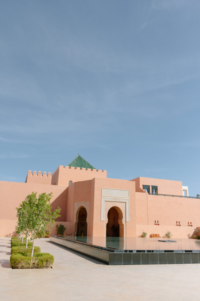 A peach-colored building with arched doorways and a green-tiled roof is reflected in a shallow pool, surrounded by neatly trimmed shrubs under a clear blue sky.