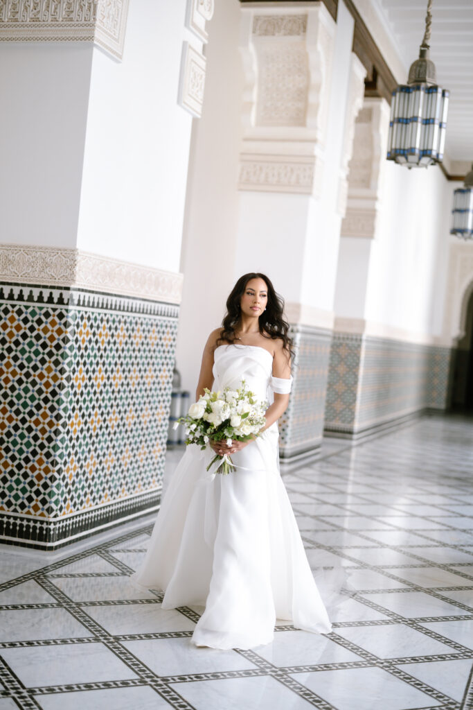 A bride in an elegant off-shoulder white gown holds a bouquet of white and green flowers, standing in a hallway with intricate mosaic tiles and ornate architectural details.