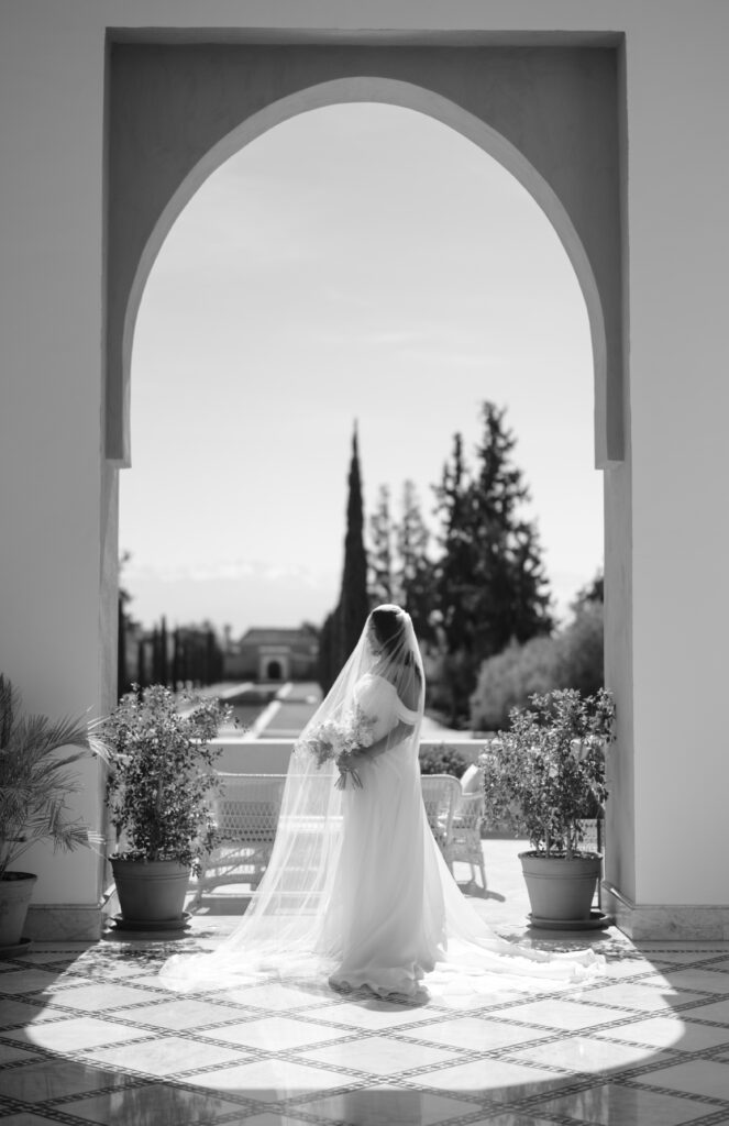 A bride in a flowing gown and veil stands under a large arched doorway, facing a sunlit garden with trees and distant buildings, framed by potted plants on a tiled floor.