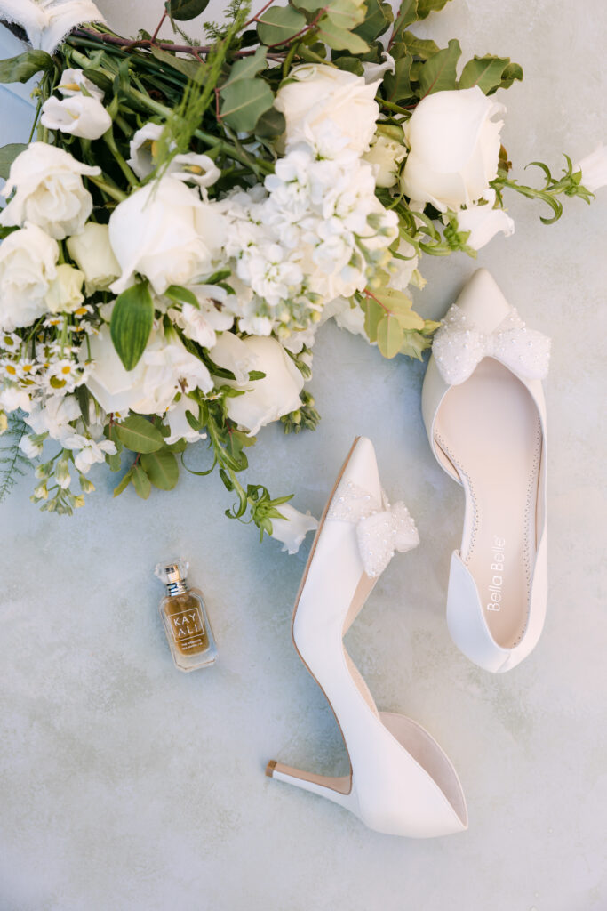 A pair of white high-heeled shoes with decorative bows, a bouquet of white flowers, and a small bottle of perfume are arranged on a light-colored surface.