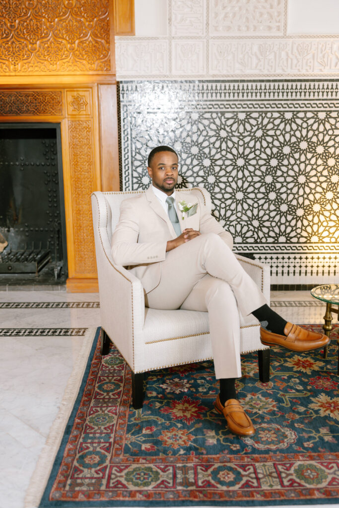A man in a light beige suit sits confidently in an armchair, legs crossed, in an elegant room with ornate black-and-white wall tiles, a decorative fireplace, and a colorful patterned rug.