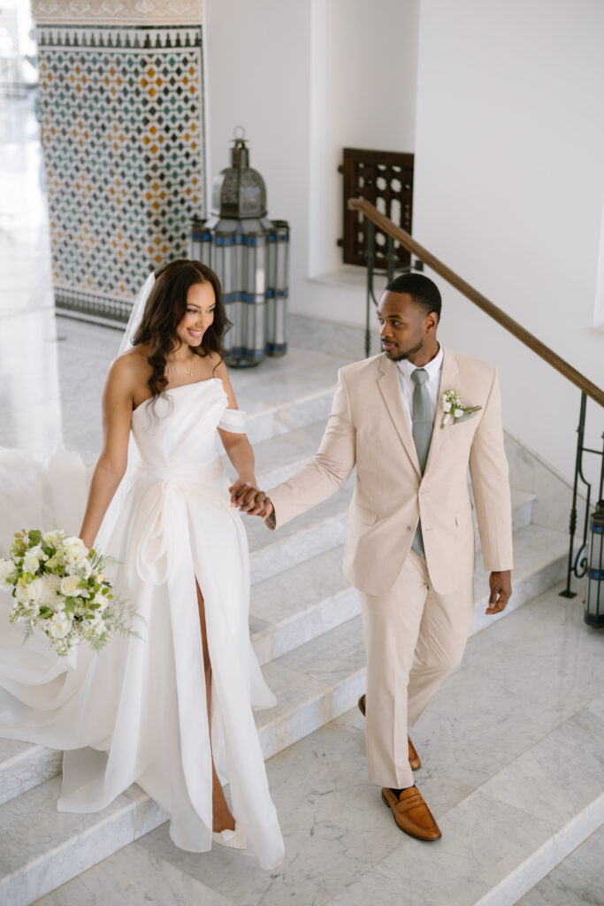 A bride in a white dress and a groom in a beige suit hold hands while walking down marble stairs, smiling at each other. The bride carries a white bouquet; patterned tiles and lanterns decorate the background.