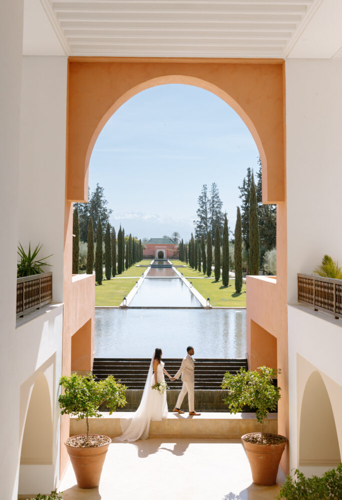 A bride and groom stand hand in hand in an elegant arched corridor, overlooking a long reflecting pool lined with tall trees, with mountains visible in the distance under a clear blue sky.