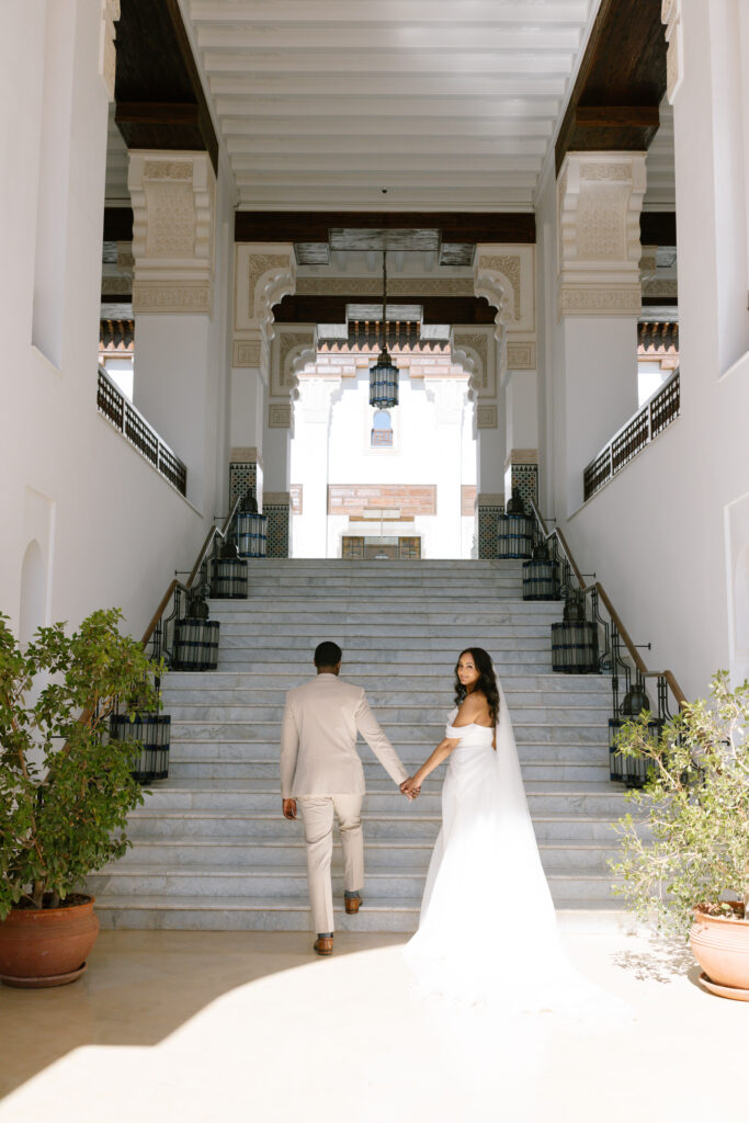 A bride and groom in elegant attire hold hands and ascend a grand marble staircase in a sunlit, ornate building with white walls, arches, and large potted plants.