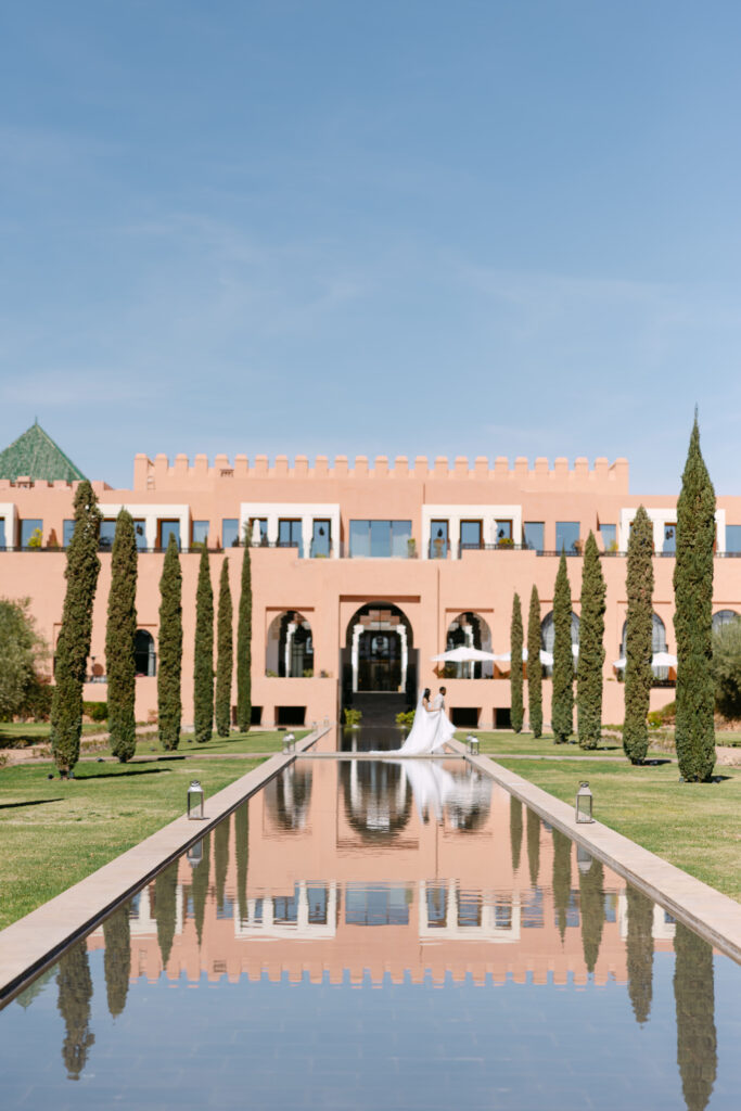 A bride and groom stand together at the edge of a long reflective pool, with a grand pink building, tall cypress trees, and a clear blue sky in the background.