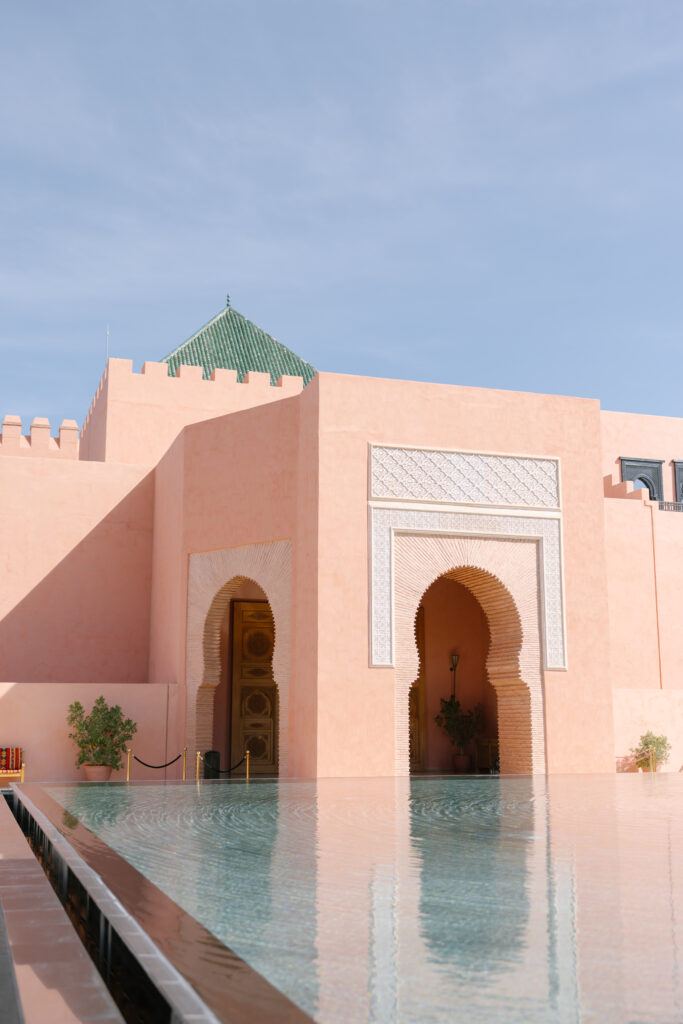 A peach-colored building with Moorish architectural features, including a large arched doorway and intricate white carvings, stands next to a reflective pool under a clear blue sky.