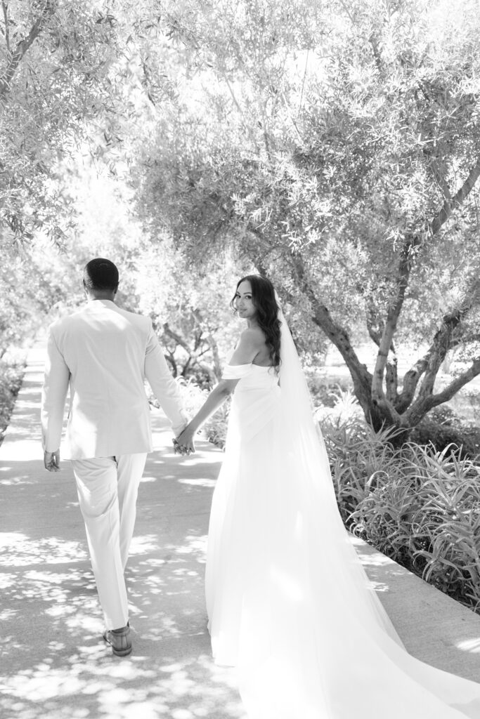 A bride and groom walk hand in hand down a sunlit path lined with trees. The bride, in a flowing white dress, looks back and smiles, while the groom, in a light suit, walks beside her. The scene is bright and serene.