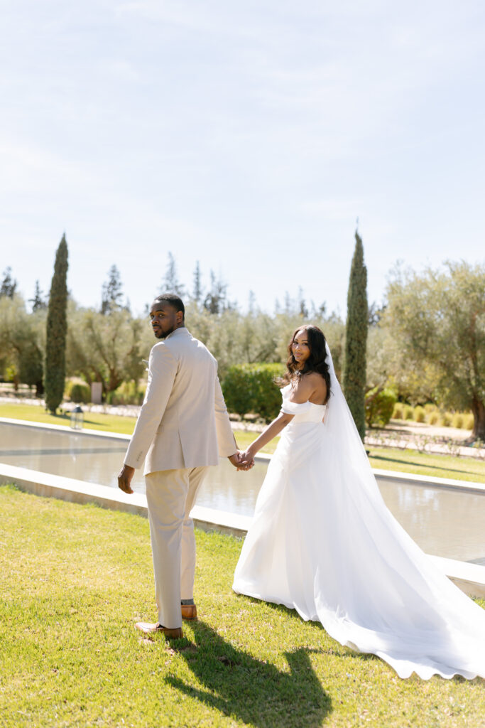 A bride in a white gown and veil holds hands with a groom in a beige suit as they walk on grass beside a reflecting pool, looking back over their shoulders on a sunny day. Tall trees and greenery are in the background.