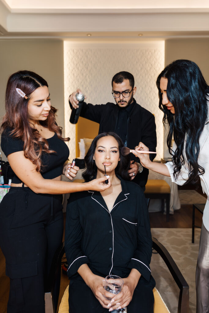 A woman in black pajamas sits on a chair holding a glass of water while three people style her; one applies lipstick, another uses a makeup brush, and a third sprays her hair.