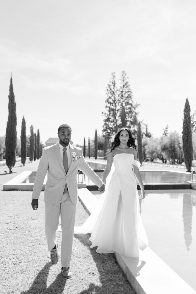 A bride and groom walk hand in hand outdoors by a long reflecting pool, surrounded by tall, narrow trees. The bride wears an off-the-shoulder gown and the groom is in a light suit. The scene is bright and elegant.