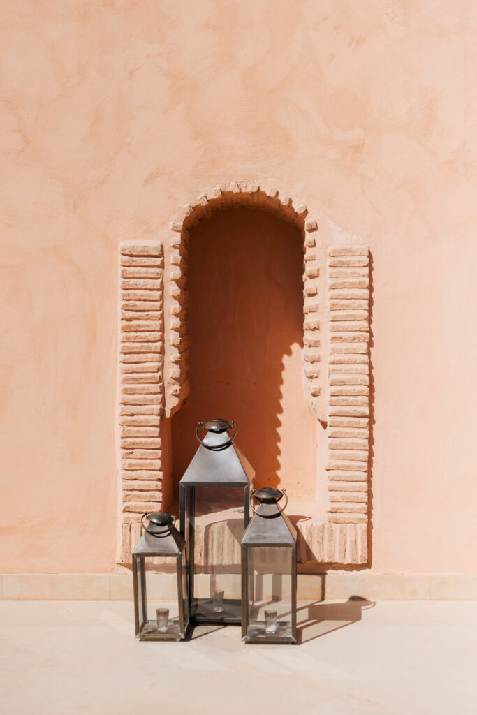 Three metal lanterns with glass sides are placed on the ground in front of a peach-colored stucco wall featuring a small arched alcove with decorative brick detailing.