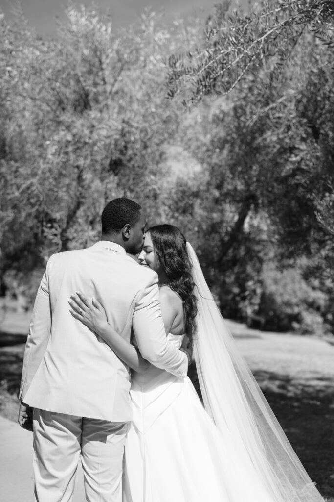 A bride and groom embrace outdoors under trees. The groom kisses the bride’s forehead as she smiles, her long veil draped behind her. The image is in black and white.