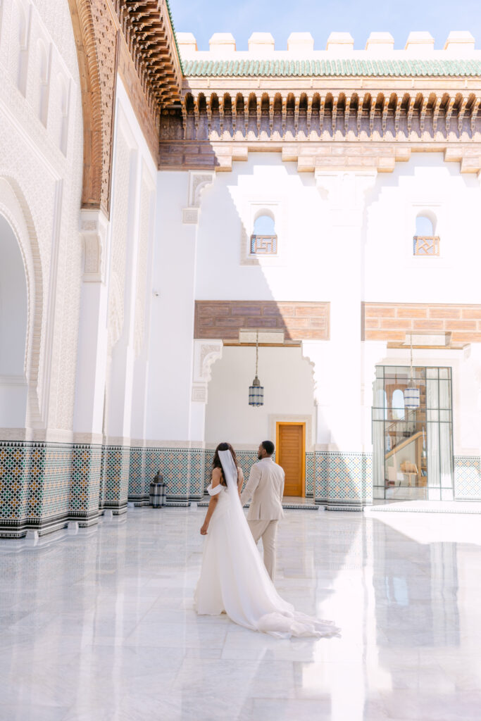 A bride and groom stand together in a sunlit, elegant courtyard with white walls, intricate arches, mosaic tiles, and wooden architectural details. The bride wears a long, flowing gown and veil.