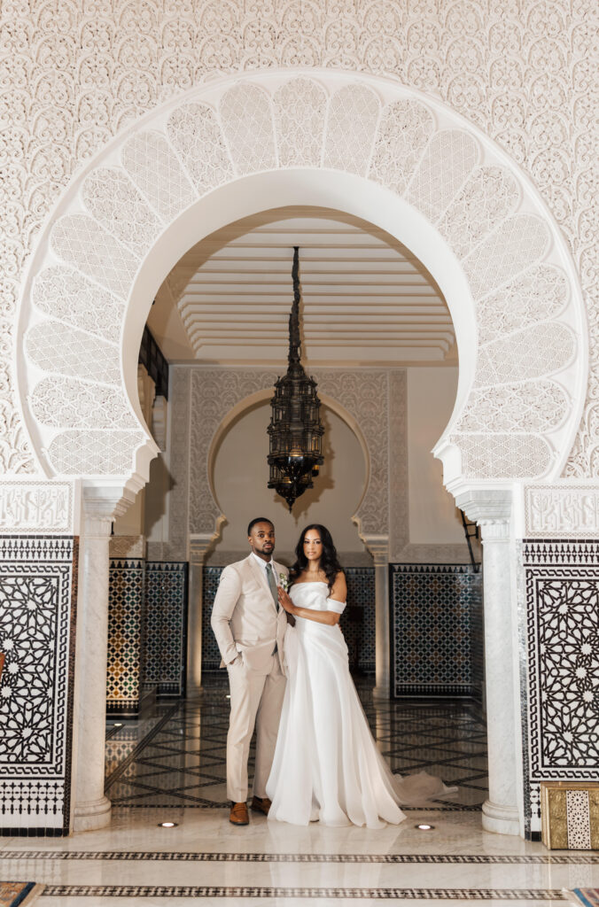A bride in a white off-shoulder gown and a groom in a beige suit stand together under an ornate, arched doorway with intricate patterns and a hanging lantern in an elegant, tiled hall.