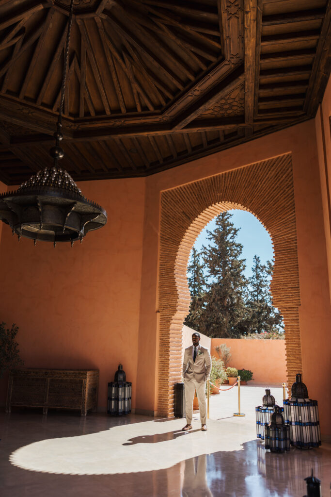 A man in a beige suit stands under an ornate wooden ceiling and beside a large arched doorway with intricate brickwork, with sunlight streaming in and casting shadows on the floor. Lanterns and trees are visible outside.