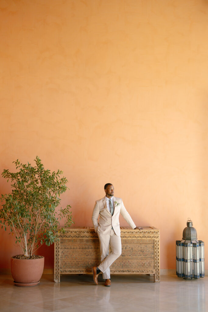 A man in a light beige suit leans against an ornate wooden chest, standing on a polished floor in front of a peach-colored wall. A potted plant and decorative lantern sit nearby.